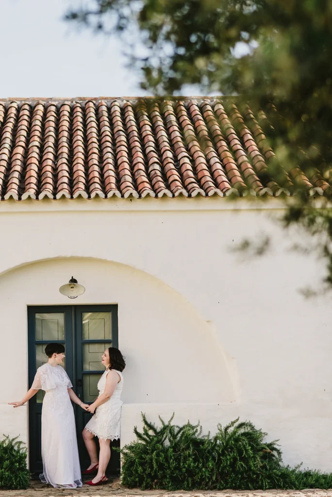 A newlywed couple in white wedding dresses shares a joyful moment outside a Mediterranean-style building with a tiled roof and an arched doorway. Holding hands, they smile warmly at each other against a minimalist white wall, framed by lush greenery.