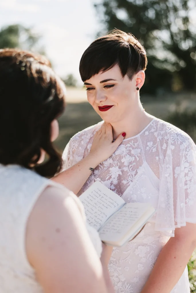 A bride in a delicate lace dress gazes at her partner with emotion as she listens to wedding vows. A handwritten notebook is held between them, capturing the raw and heartfelt moment of their intimate outdoor ceremony.