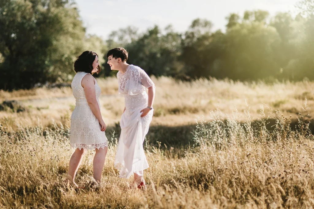 A newlywed couple in white lace dresses shares a joyful laugh in a sunlit field during golden hour. The soft light and golden grass create a romantic and carefree atmosphere as they enjoy a lighthearted moment together.