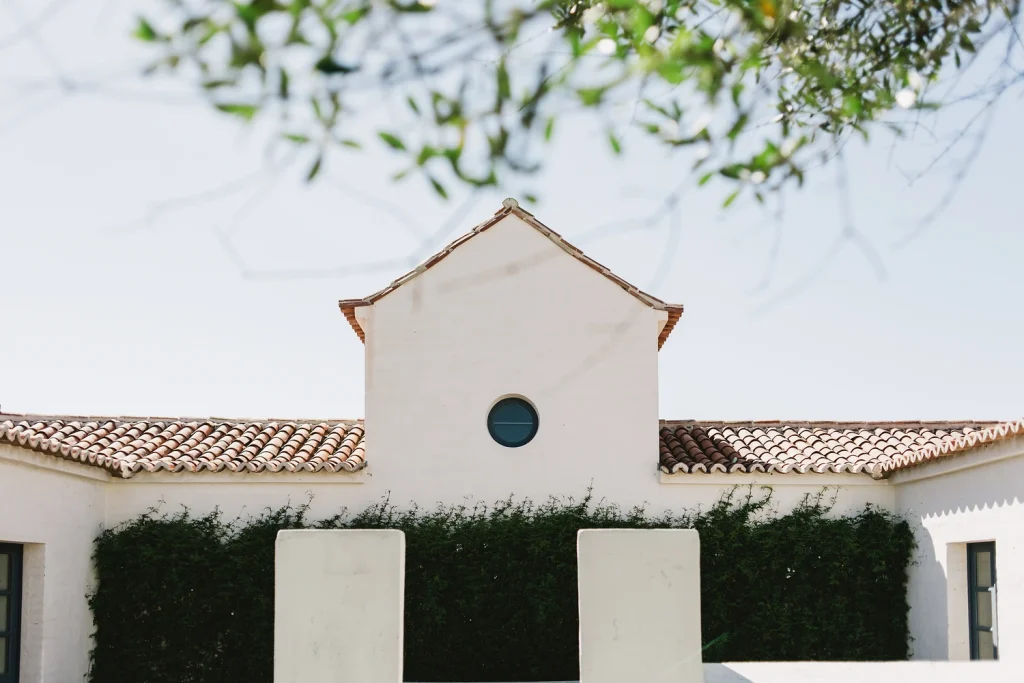 A beautifully minimalist Mediterranean-style building with white stucco walls, terracotta roof tiles, and a circular window. Green vines climb along the lower facade, while soft olive tree branches frame the top, creating a serene and elegant composition.
