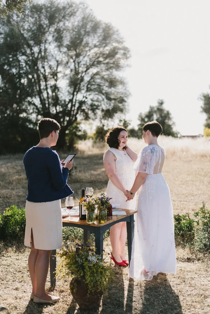 A couple in white lace dresses holds hands during an intimate outdoor wedding ceremony, surrounded by golden fields and lush trees. A friend officiates while reading from a tablet, and a rustic table with wine and flowers adds to the natural elegance of the scene.