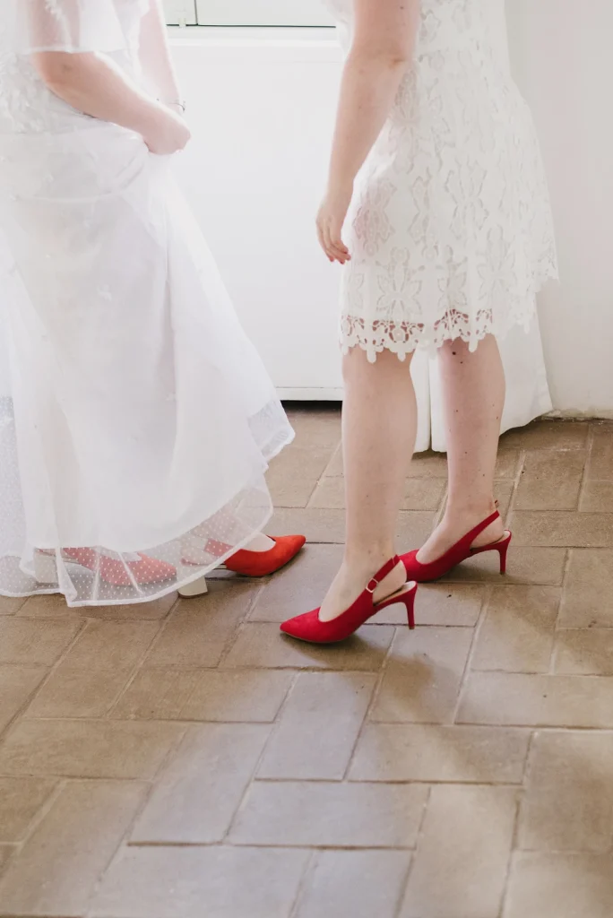 A close-up of two brides wearing white lace dresses and bold red shoes, standing on a tiled floor. The striking footwear choice adds a stylish and personal touch to their wedding attire.
