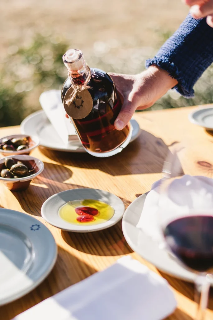 A hand pours red wine from a rustic bottle onto a wooden outdoor table set with olive oil, fresh olives, and traditional ceramic plates. The sunlight enhances the warm, inviting atmosphere of a Mediterranean-inspired dining experience.