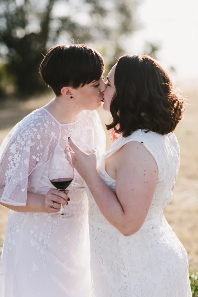 A newlywed couple in elegant white lace dresses shares a romantic kiss in a sunlit field during golden hour. One bride holds a glass of red wine, while the soft sunlight enhances the intimate and heartfelt moment.