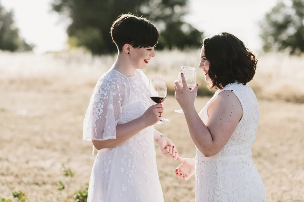 A newlywed couple in elegant white lace dresses clinks wine glasses in a golden field, sharing a joyful and intimate toast. Their hands gently intertwine, capturing a romantic and celebratory moment in the warm sunlight.