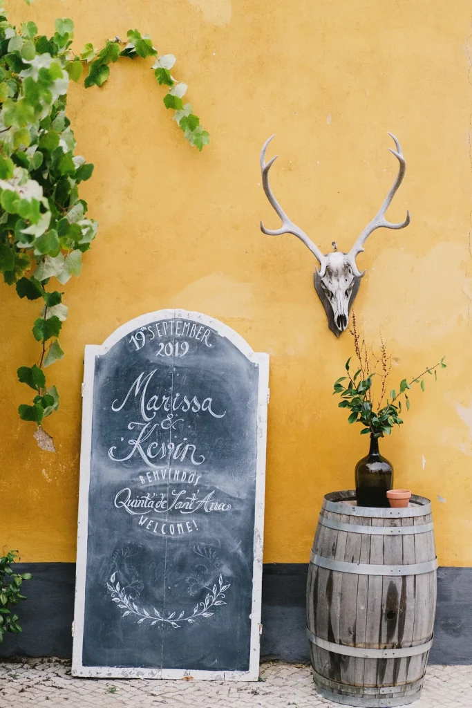 A rustic wedding welcome sign for Marissa & Kevin, dated September 19, 2019, at Quinta de Sant'Ana. The black chalkboard with elegant white calligraphy leans against a yellow stucco wall, accompanied by a wooden barrel with a glass bottle vase and a mounted deer skull with antlers. Green vines add a natural touch to the scene.