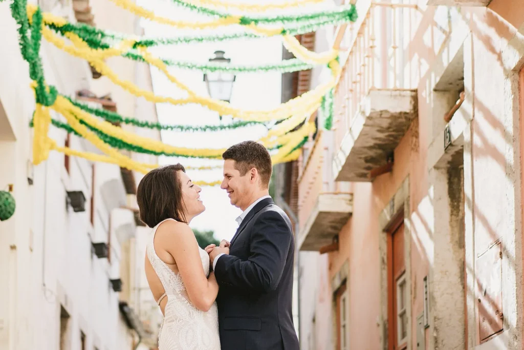A newlywed couple dances joyfully under colorful festival streamers in a charming European street.