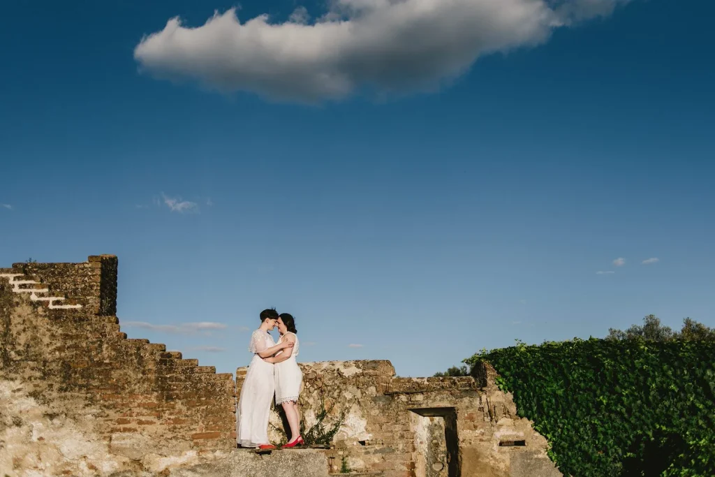 A same-sex couple kisses atop an ancient stone ruin under a vast blue sky, with a single cloud floating above them.
