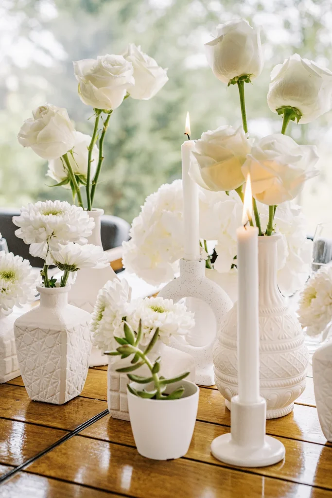 Elegant white wedding centerpiece with roses, candles, and modern ceramic vases on a wooden table.