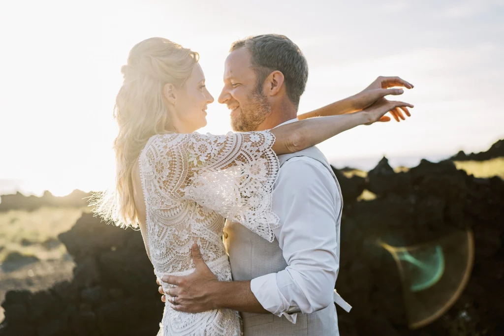 A couple embraces at sunset, the bride in a lace gown lifting her arms joyfully as they share a romantic moment.