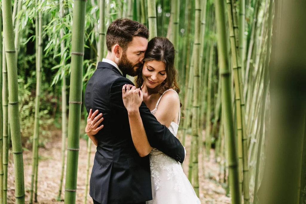 A newlywed couple embraces in a lush bamboo forest, the bride smiling while leaning into her groom.