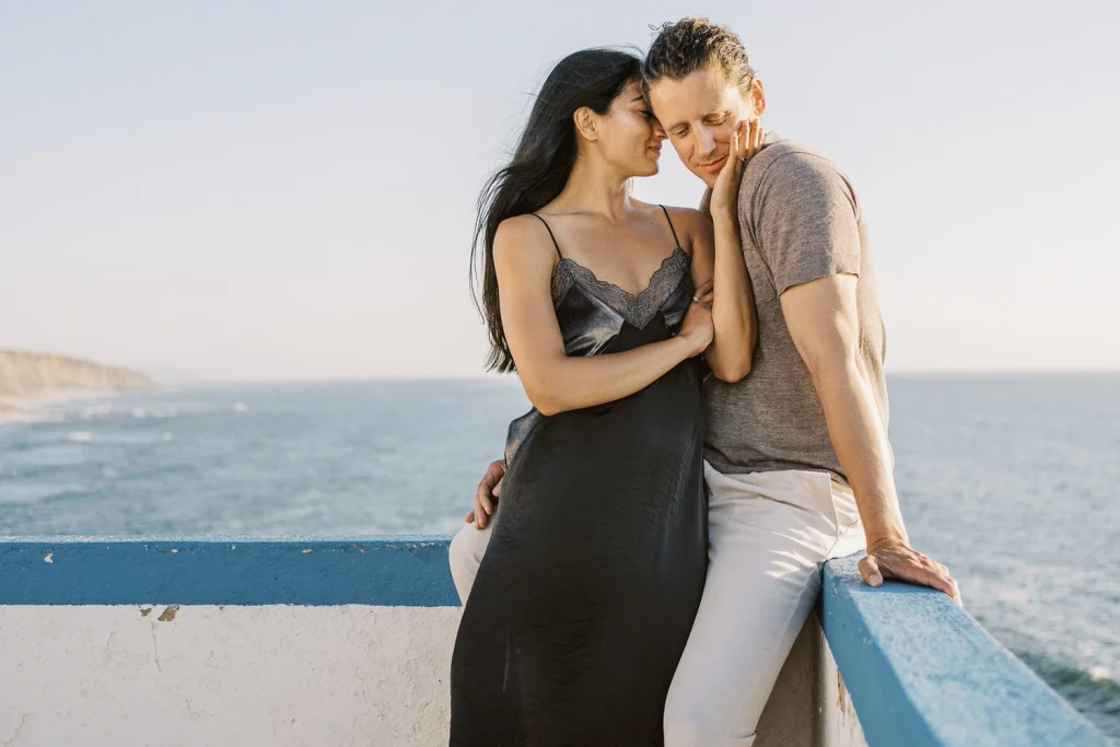 A couple leans against a blue and white wall near the ocean, sharing a quiet and intimate moment.