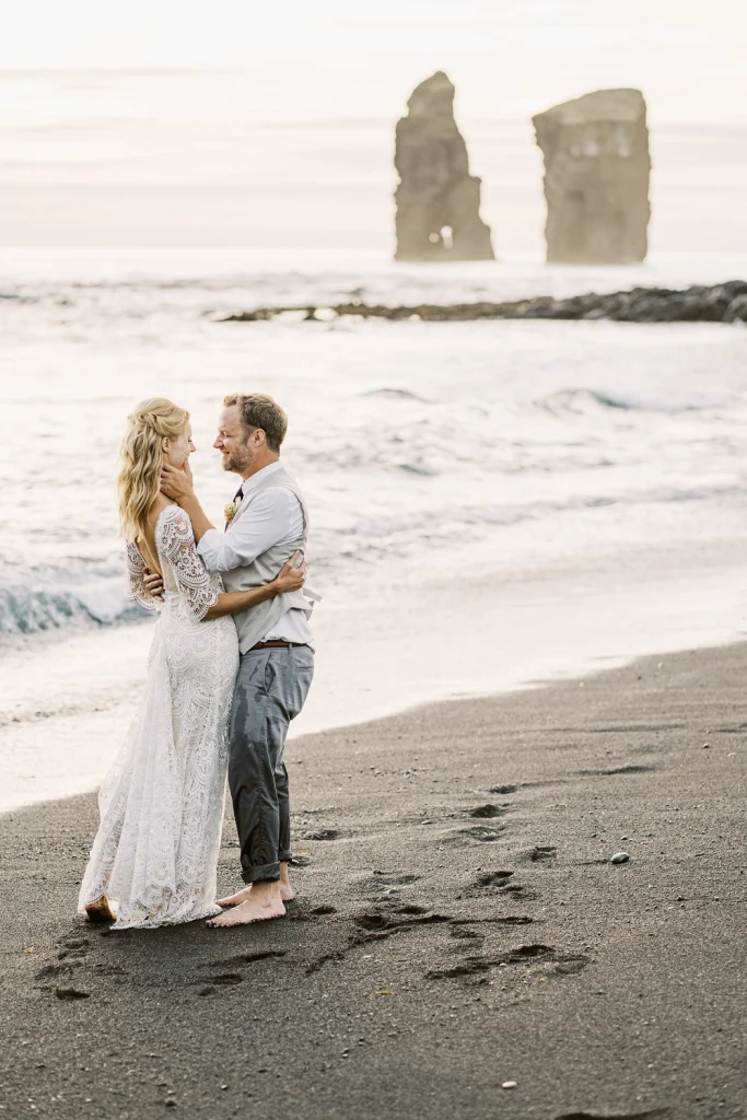 A bride and groom embrace barefoot on a black sand beach, smiling at each other as waves crash nearby, with two dramatic sea stacks rising in the ocean at sunset.