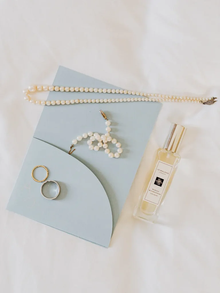 A flat-lay composition featuring a blue wedding invitation, pearl necklace, earrings, wedding rings, and a Jo Malone perfume bottle on a soft white fabric background.