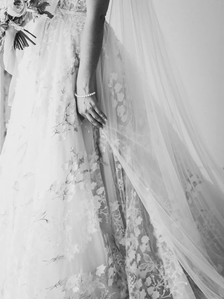 A black-and-white close-up of a bride’s hand gently lifting the lace train of her wedding dress, showcasing intricate floral embroidery and a delicate pearl bracelet.