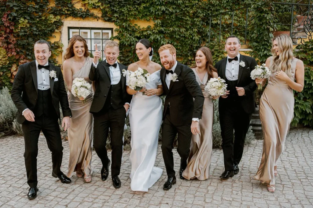 A bride and groom walk arm in arm with their wedding party, all dressed in elegant attire, laughing together.