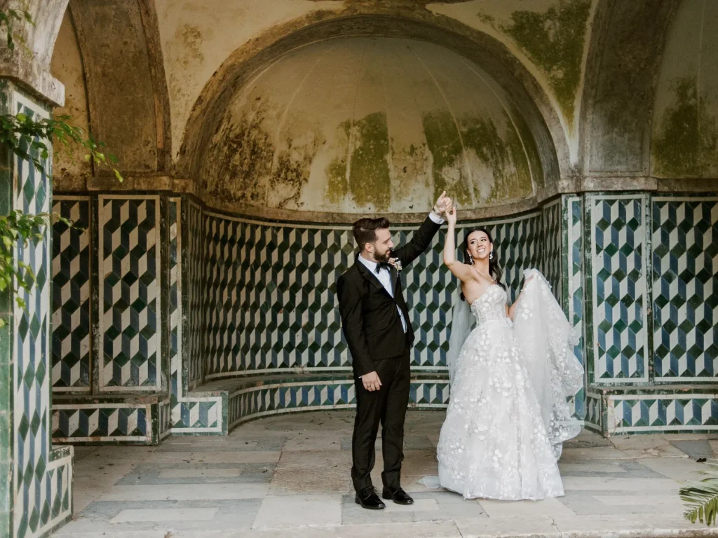 Groom in a black tuxedo twirls his bride in an elegant lace gown under an ornate, historic archway with geometric tiled walls.