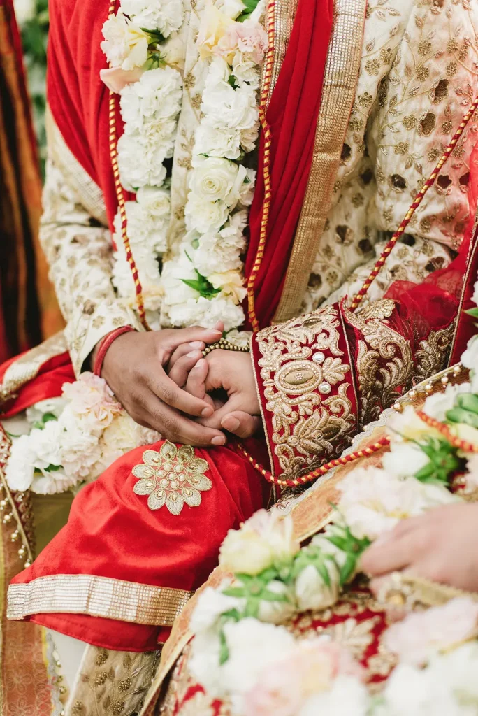 A close-up of the bride and groom’s hands gently intertwined during their Hindu wedding ceremony at Quinta de Sant’Ana, Portugal. Their red and gold embroidered attire, adorned with floral garlands, symbolizes love and commitment.