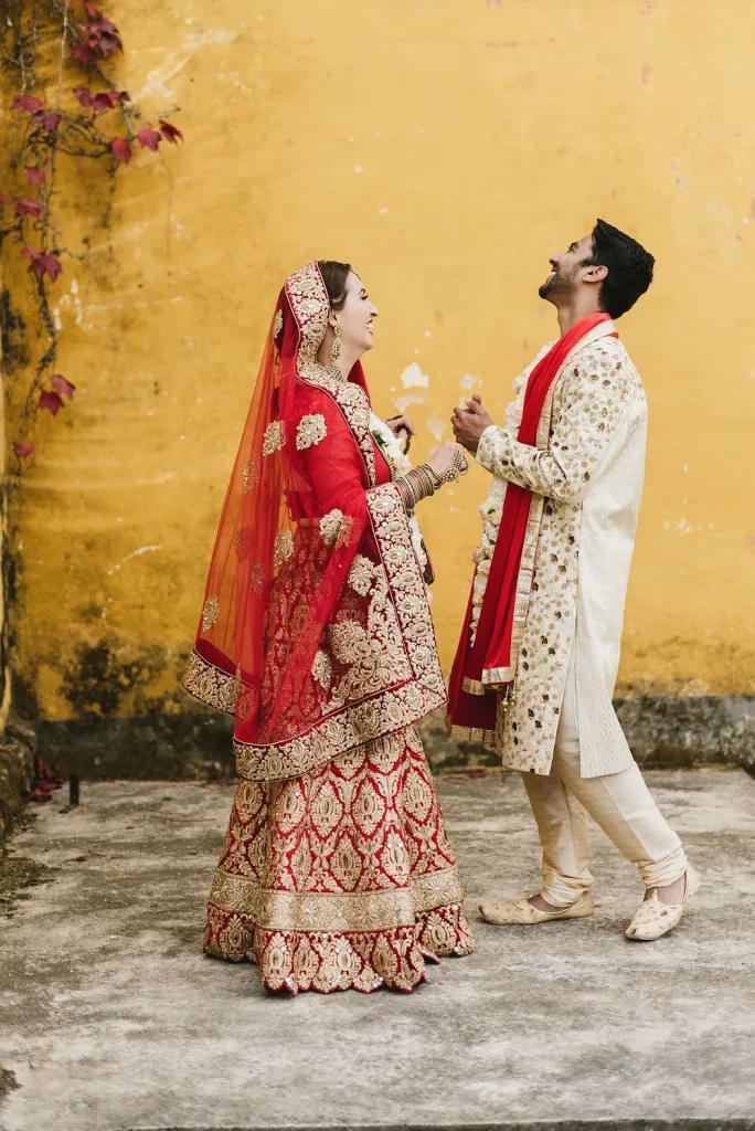 A joyful moment between the bride and groom at Quinta de Sant’Ana, Portugal. Dressed in traditional Hindu wedding attire, they share laughter against a rustic yellow wall adorned with autumnal vines.