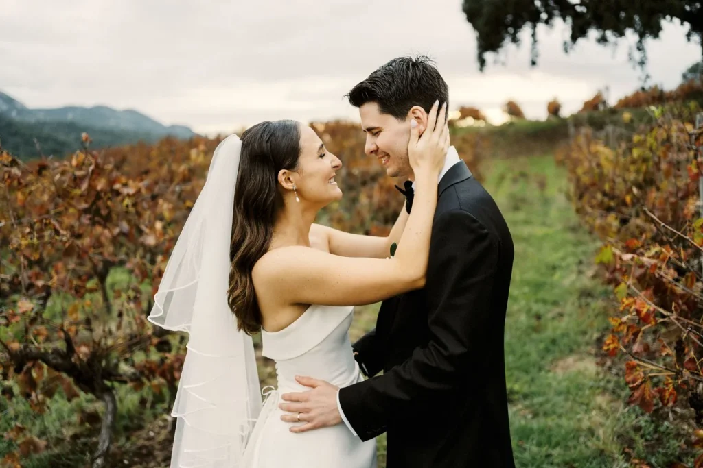 A newlywed couple embraces in a vineyard, gazing into each other’s eyes with joy.