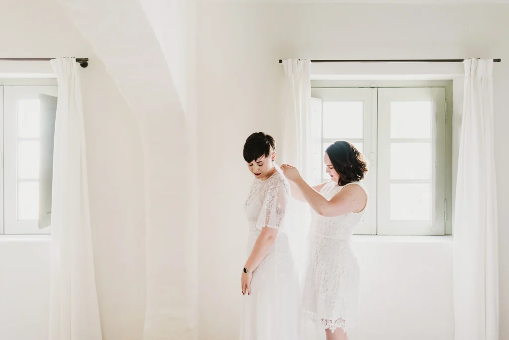 A bride fastening her partner’s wedding dress in a bright, minimalist room with natural light in Portugal.