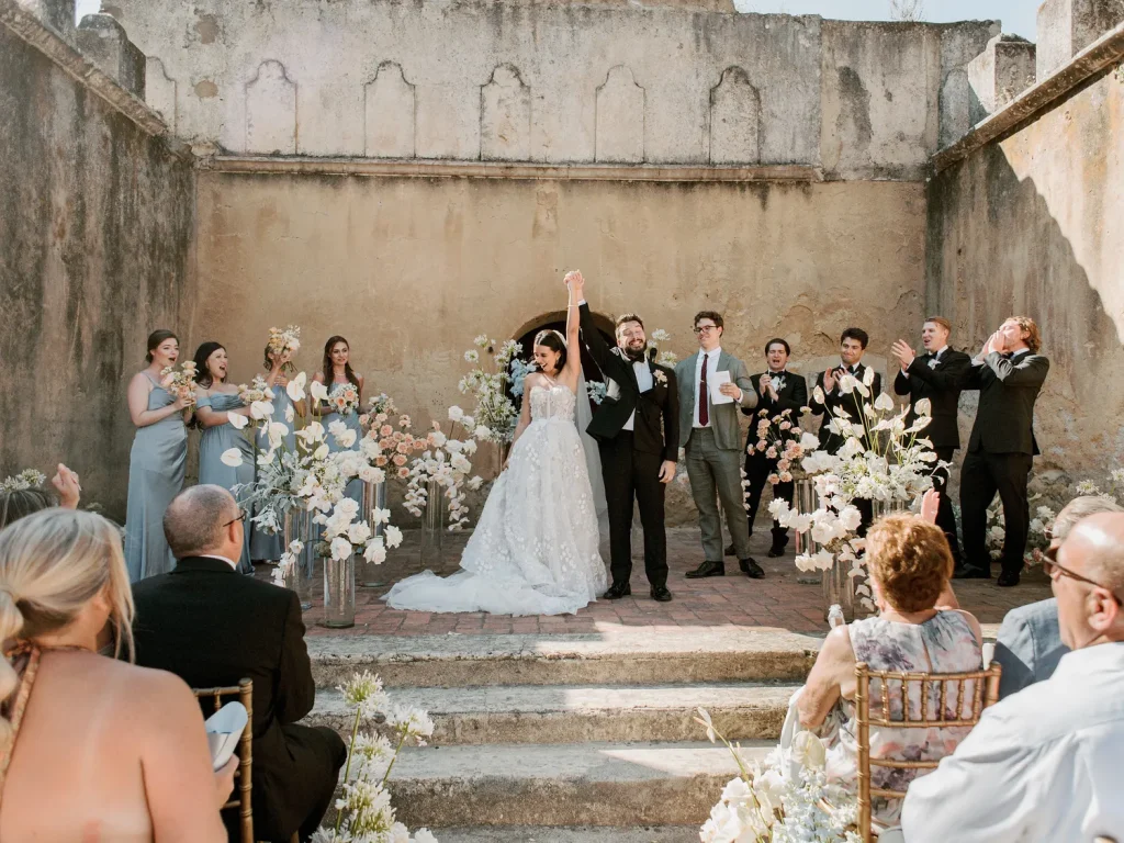 A newlywed couple raises their hands in celebration at the altar, surrounded by bridesmaids, groomsmen, and cheering guests in an intimate outdoor wedding setting.