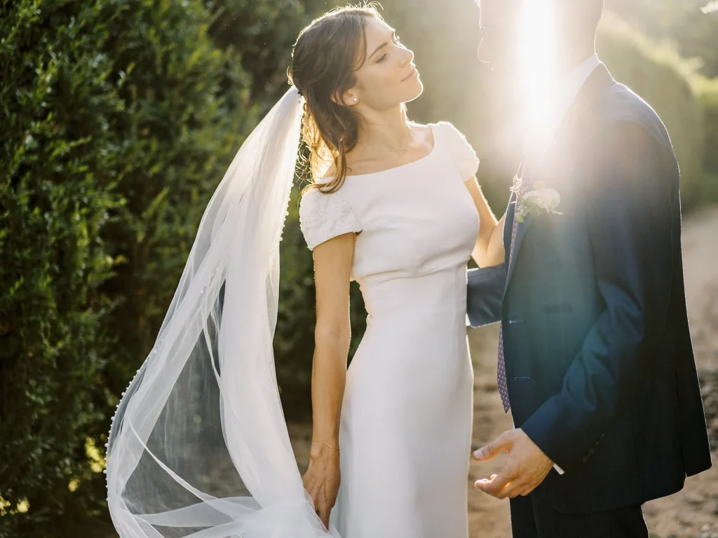 A bride gazes at her groom during golden hour, with sunlight streaming between them, creating a dreamy and romantic glow.