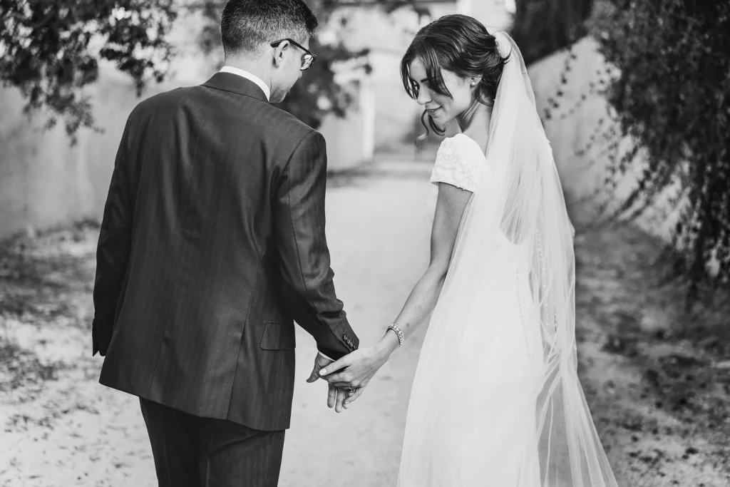 Black and white photograph of a bride and groom walking hand in hand down a rustic path, capturing a quiet and intimate newlywed moment.