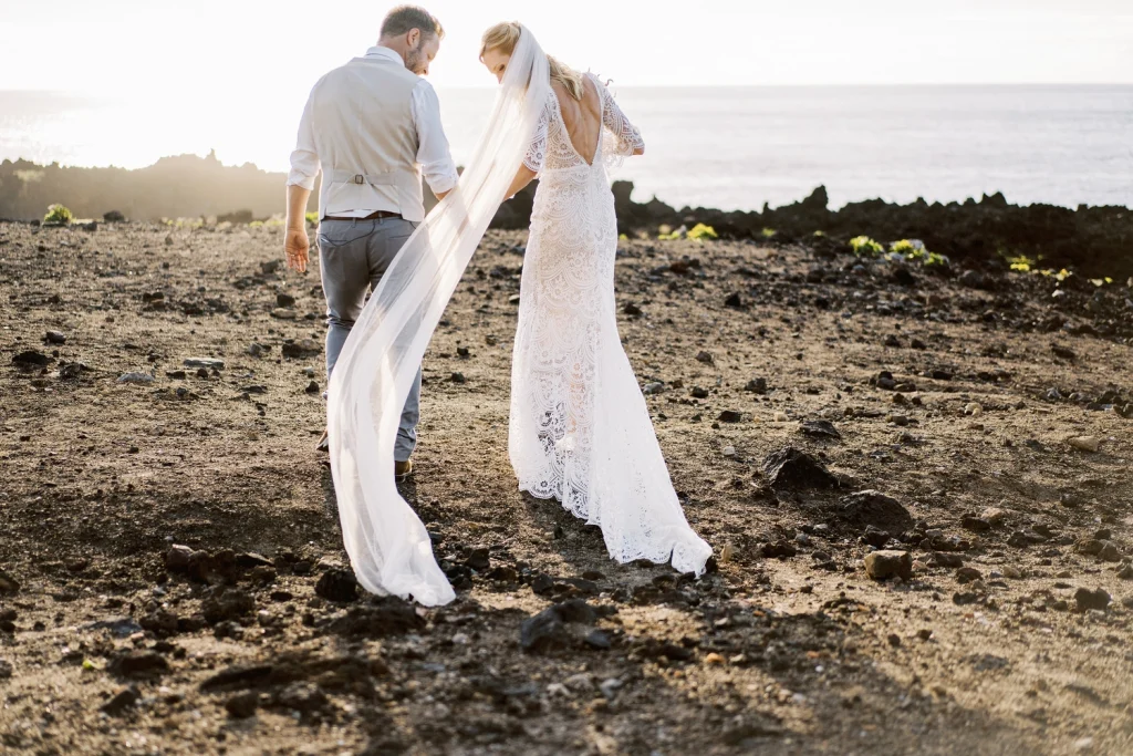 A bride and groom walk hand in hand across a rugged volcanic landscape, with the bride’s delicate lace dress and flowing veil trailing behind her in the golden sunset light.