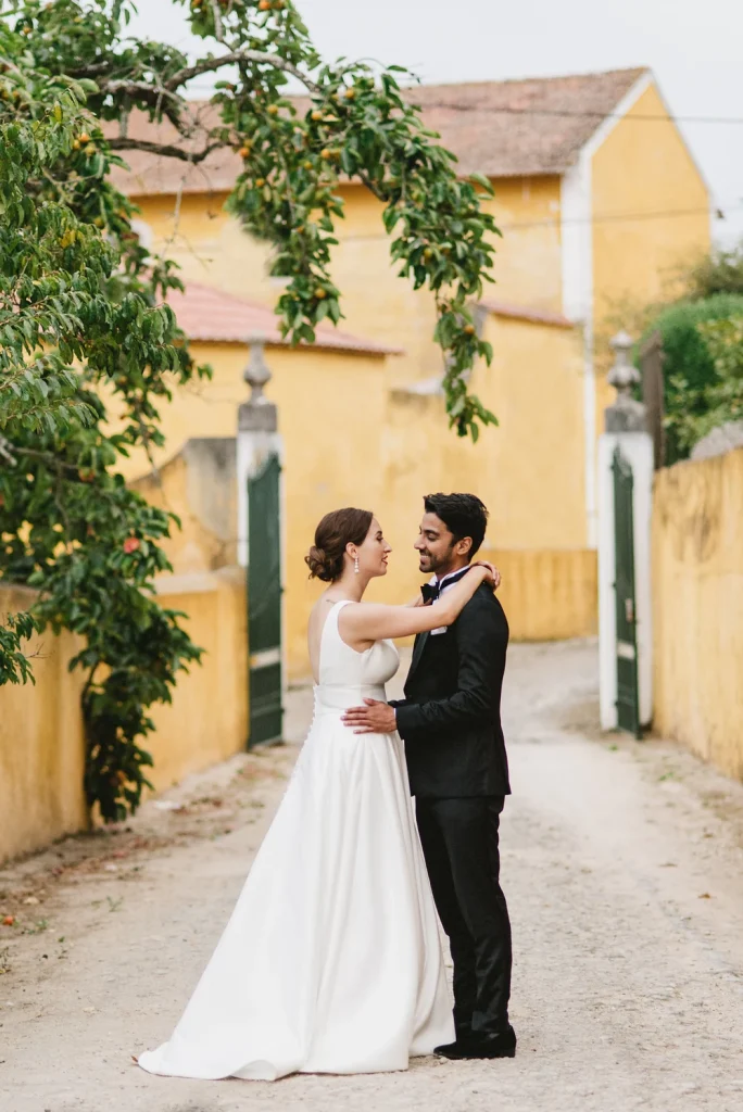 A newlywed couple embraces in a charming cobblestone pathway at Quinta de Sant’Ana, Portugal. The bride, in a flowing white gown, and the groom, in a classic black tuxedo, share a joyful moment against a backdrop of yellow-washed walls, green gates, and lush overhanging branches. The rustic setting adds a romantic and timeless feel to the scene.
