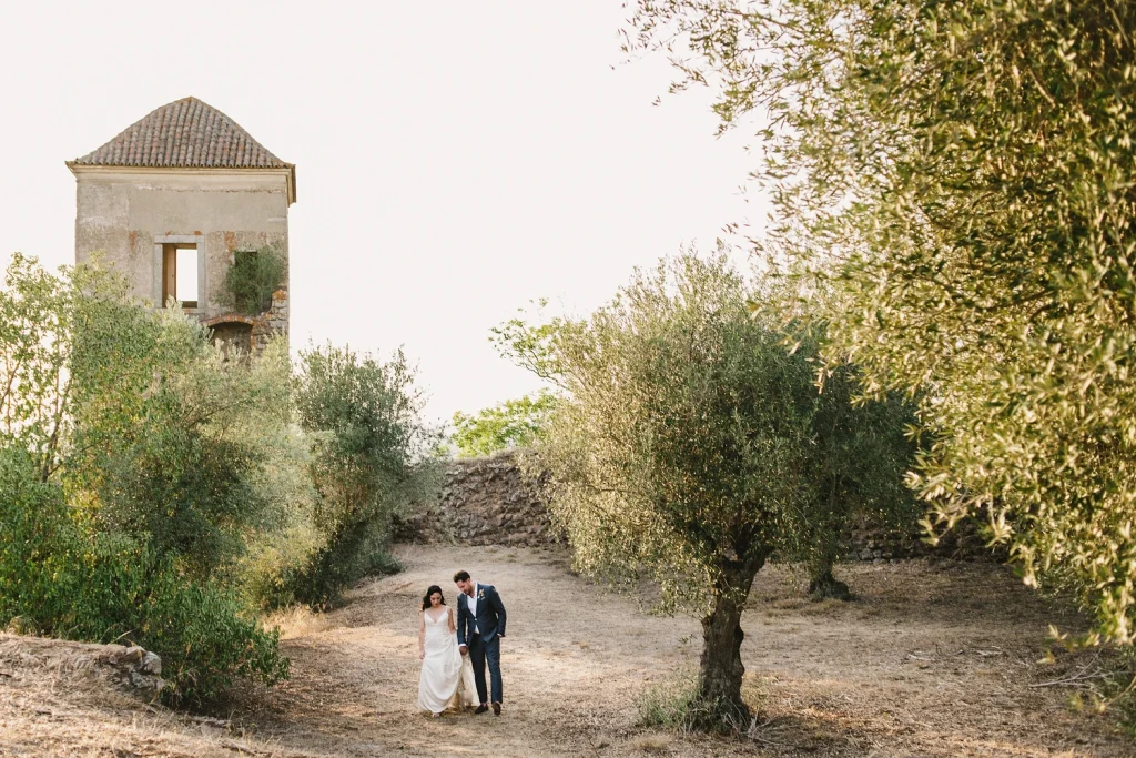 A newlywed couple walks hand in hand through an olive grove near a historic stone tower, bathed in golden sunset light.