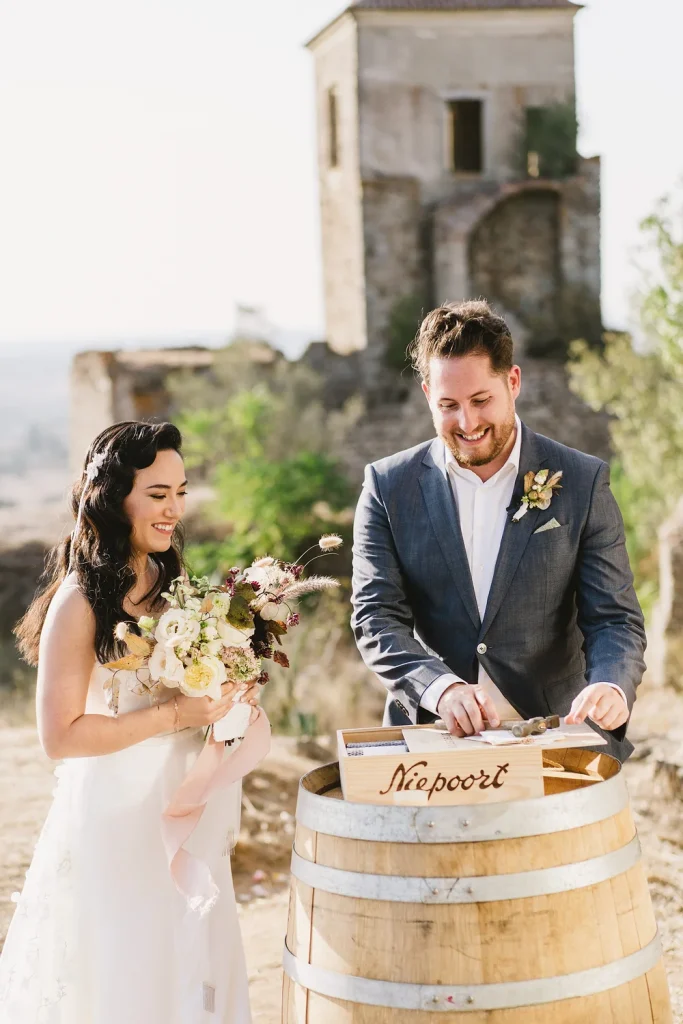 A smiling bride and groom participate in a wine box ceremony, sealing a wooden wine crate on a rustic barrel with a historic ruin in the background.
