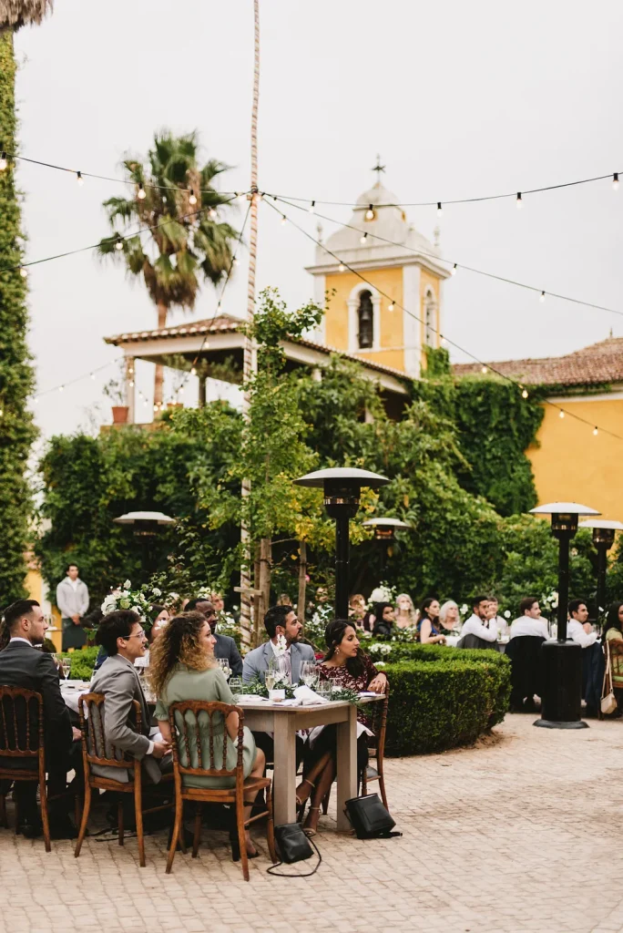 Wedding guests enjoy an intimate dinner under string lights in the courtyard of Quinta de Sant’Ana, Portugal. The elegant outdoor setting is surrounded by lush greenery, historic architecture, and a warm, inviting atmosphere.