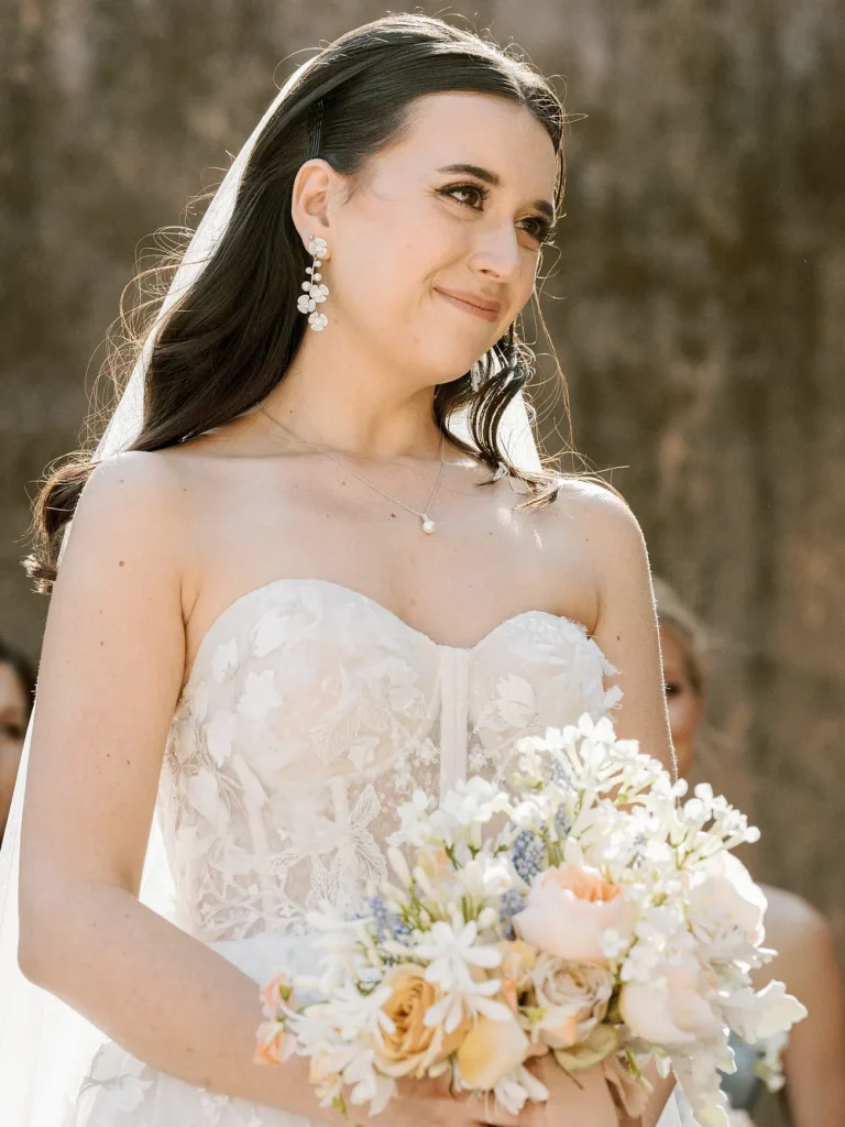 A bride smiles while holding a bouquet of pastel-colored flowers during her wedding ceremony.