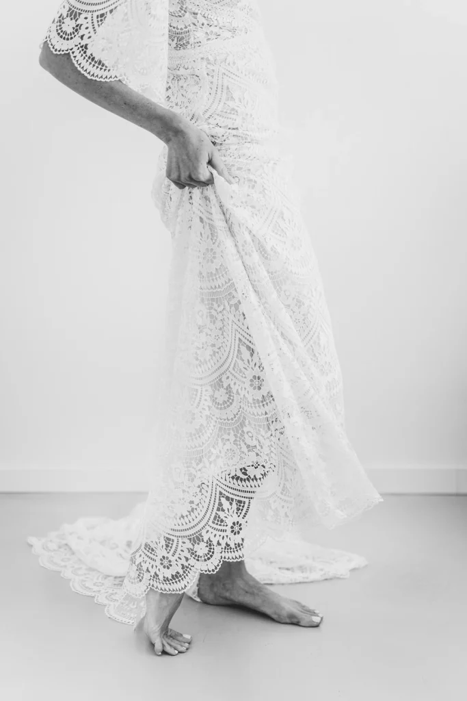 Black and white close-up of the bride lifting the hem of her lace wedding dress, revealing bare feet.