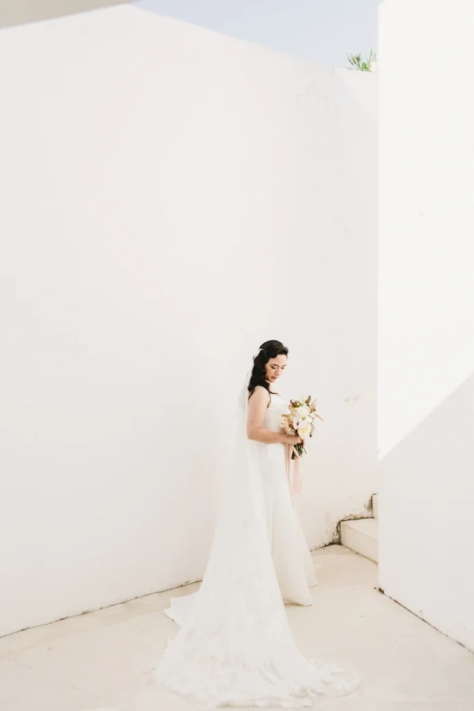 Bride standing in a modern, minimalist white space, holding a bouquet and looking down softly.