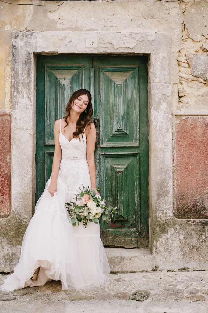 A bride in a lace wedding gown stands in front of an old green wooden door, holding a bouquet and smiling softly.