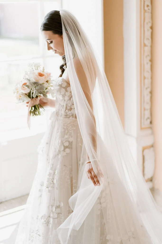 A bride holding a bouquet walks near a window, her long veil cascading behind her.