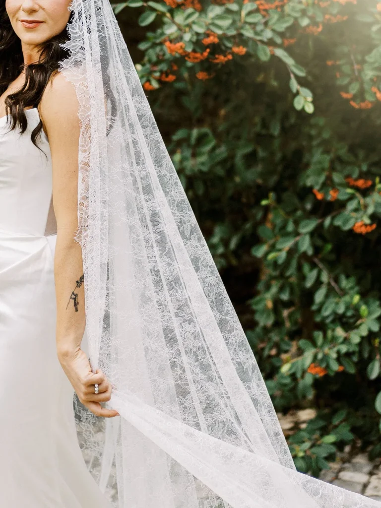 A close-up of a delicate lace wedding veil draped over the bride’s arm, with green foliage in the background.