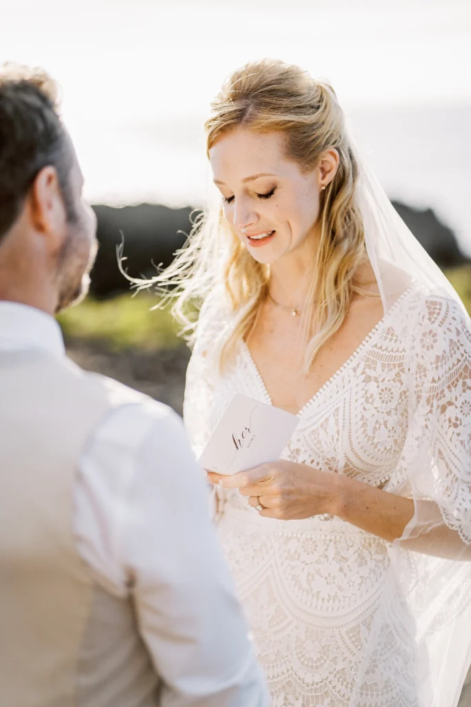 A bride with golden hair and a delicate lace dress reads her wedding vows from a small booklet, smiling softly as she stands in the golden sunlight.
