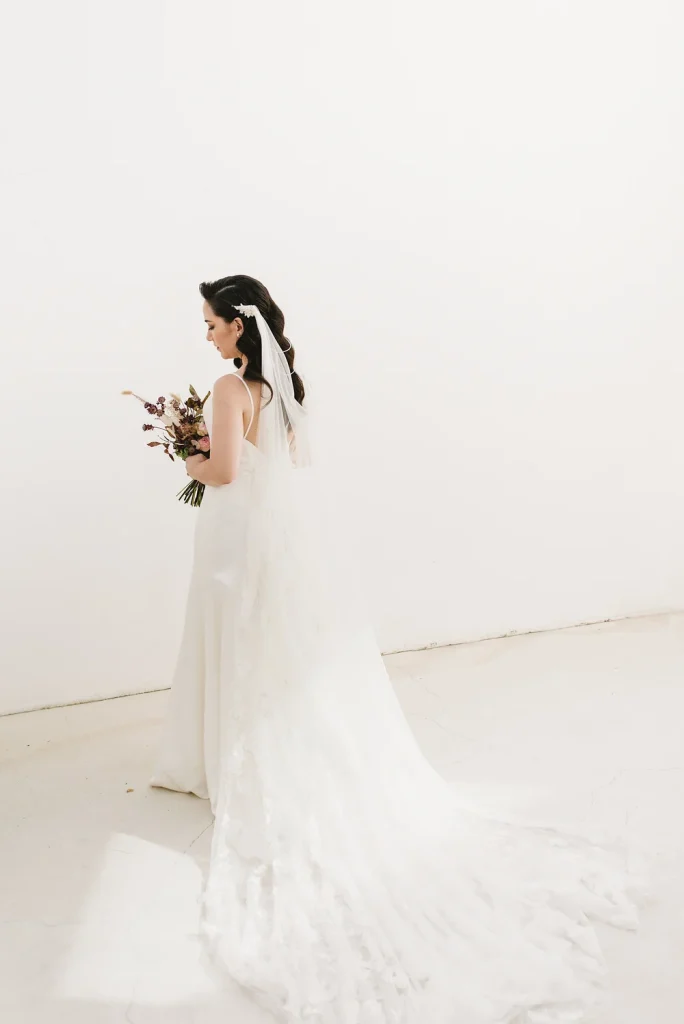 Bride in a flowing wedding gown with a long veil, holding a bouquet against a minimalist white wall.