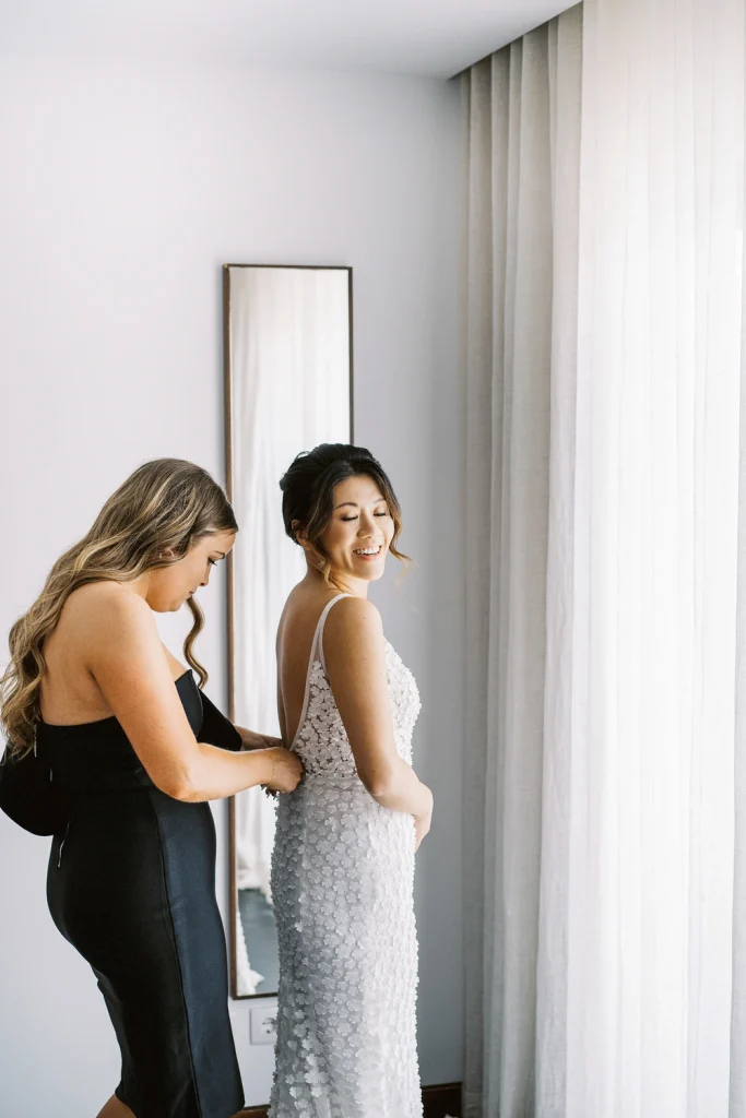 Bridesmaid helping the bride zip up her white wedding dress near a bright window.