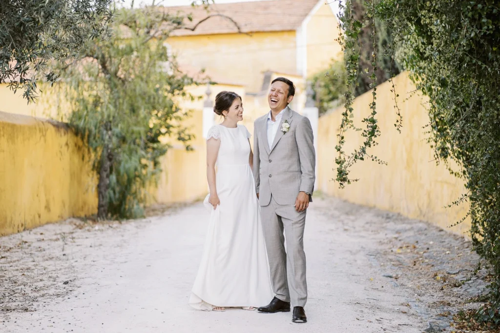 A bride in a simple white dress and a groom in a grey suit laugh together while walking down a sunlit alleyway.