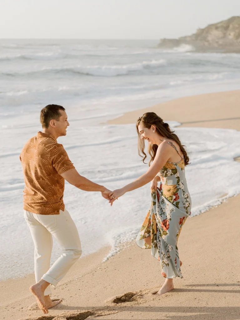 A couple playfully holds hands while walking along the shoreline with waves washing over their feet.