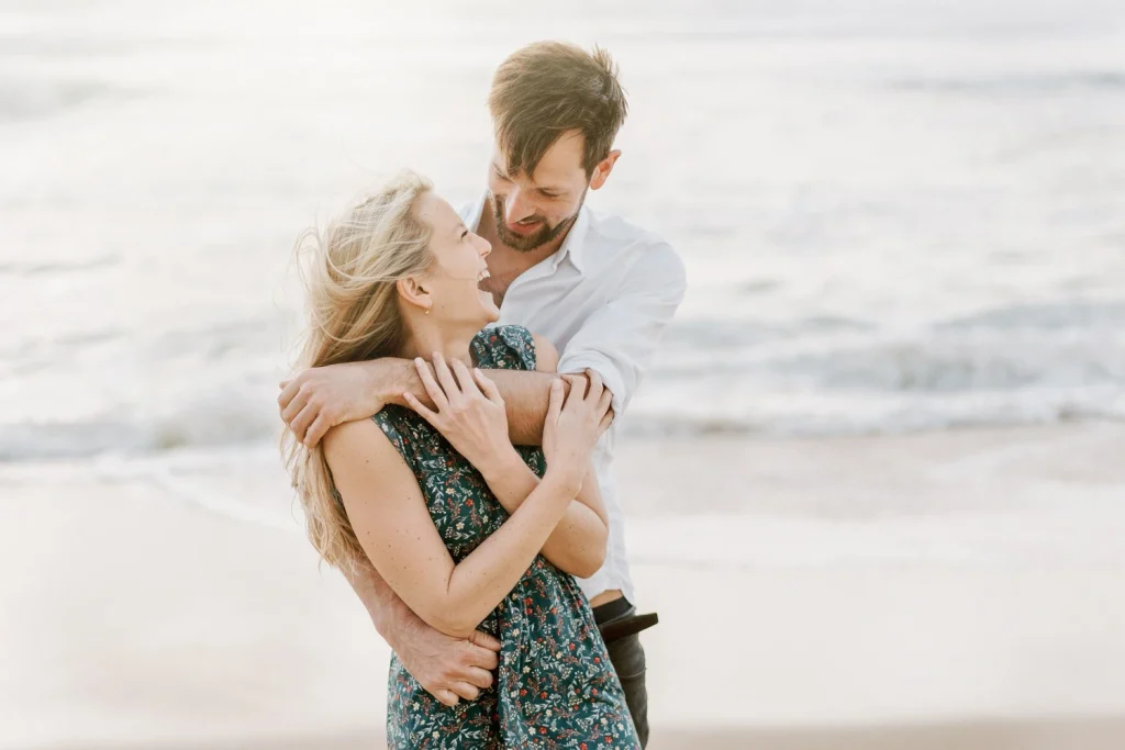 A man embraces his partner from behind as they share a joyful moment near the beach.