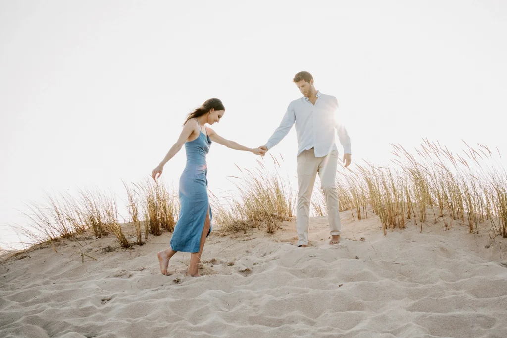 A couple walks hand in hand through soft sand dunes, bathed in golden sunlight.