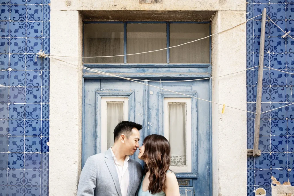 A couple leans close together in front of a rustic blue door with clotheslines above them, capturing a romantic, timeless moment.