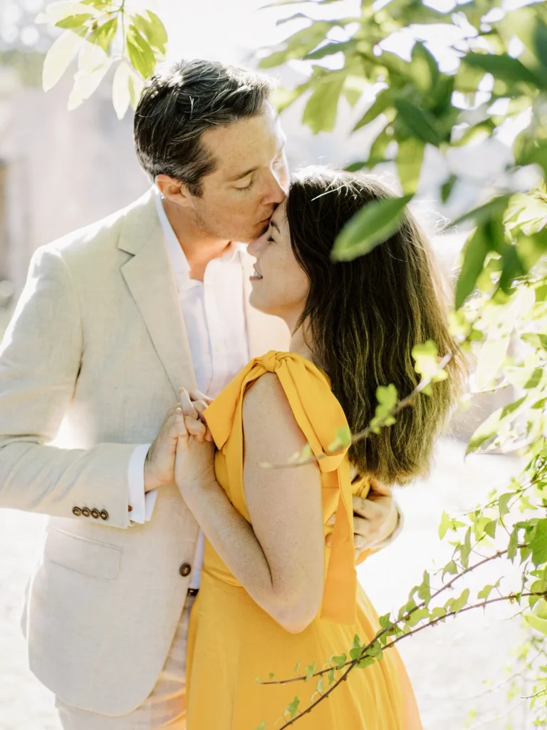 A couple shares an intimate moment under dappled sunlight, as the man gently kisses the woman’s forehead.