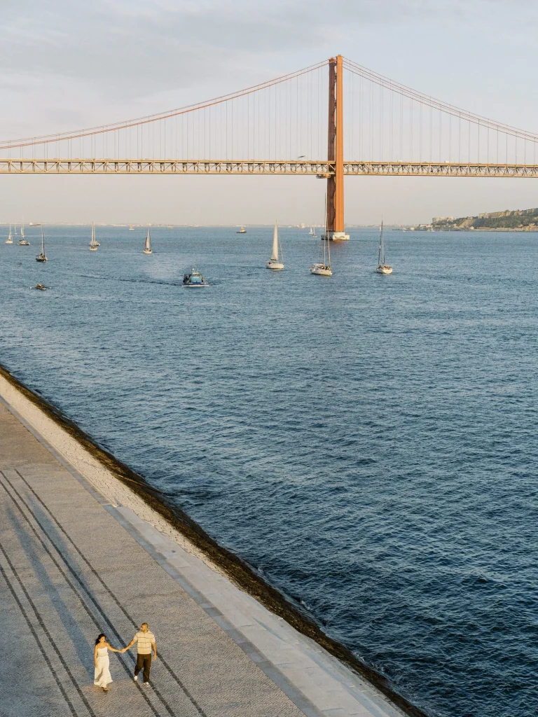 A view of the 25 de Abril Bridge spanning the Tagus River in Lisbon, with sailboats gliding across the water.