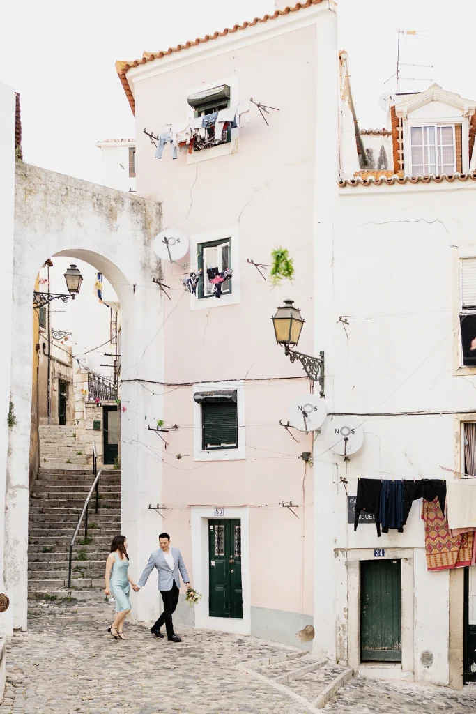 A couple walks up a stone staircase in a historic European neighborhood with pastel-colored buildings and hanging laundry.
